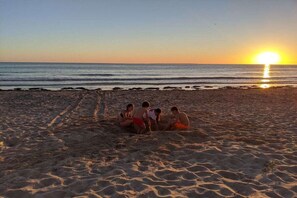 A beach scene with a group of people sitting on the sand near the water during sunset. - Holiday home in Vesly with swimming pool (Vesly)