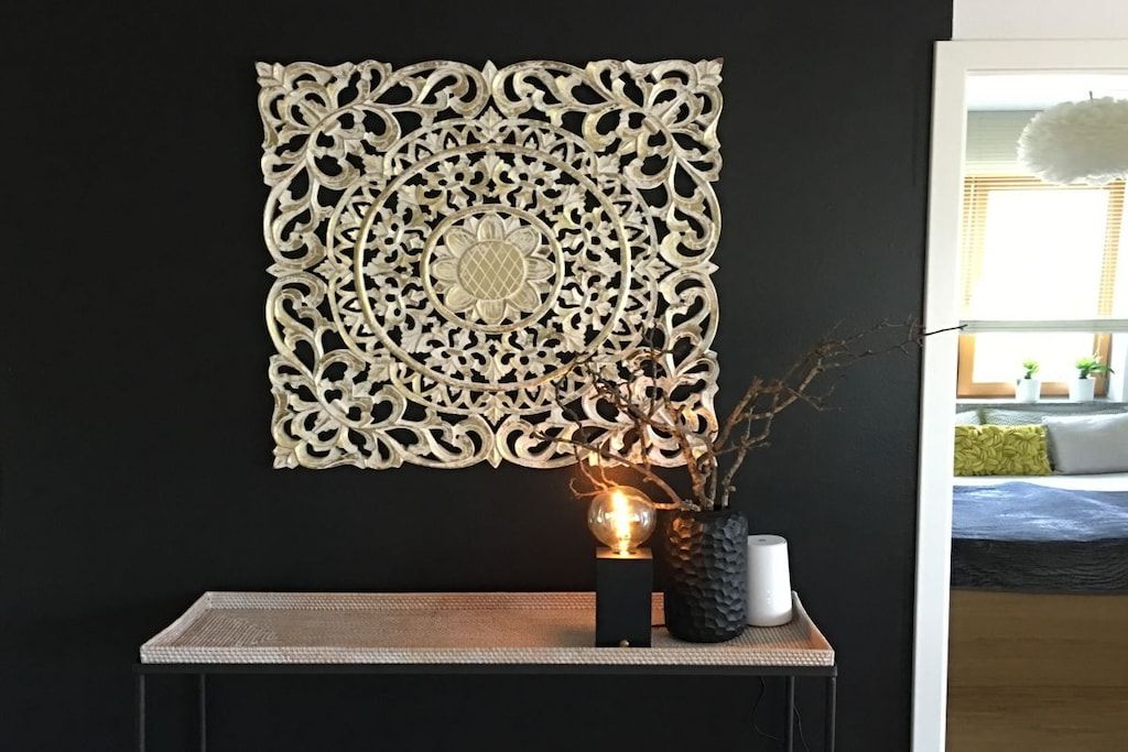 A decorative wall piece with intricate patterns, a small table with a vase and a spherical object, and a glimpse of a room with a bed and a window.