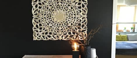 A decorative wall piece with intricate patterns, a small table with a vase and a spherical object, and a glimpse of a room with a bed and a window.
