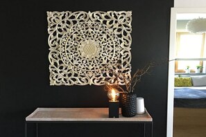 A decorative wall piece with intricate patterns, a small table with a vase and a spherical object, and a glimpse of a room with a bed and a window. - Holiday apartment with 2 bedrooms (Überlingen)