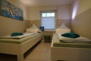 A room with two single beds, a small stool, a window with blinds, and a painting on the wall. - Holiday apartment with 2 bedrooms (Norddeich)