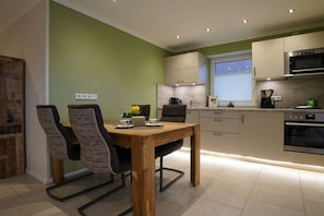 A modern kitchen with a dining area, featuring a wooden table and chairs, a built-in oven, and a window with blinds. - Holiday apartment with 2 bedrooms (Norddeich)
