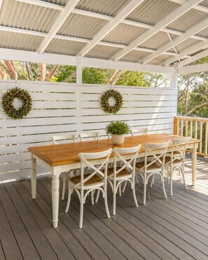 Een houten terras buiten, met een witte pergola, een eettafel met stoelen en twee potplanten.