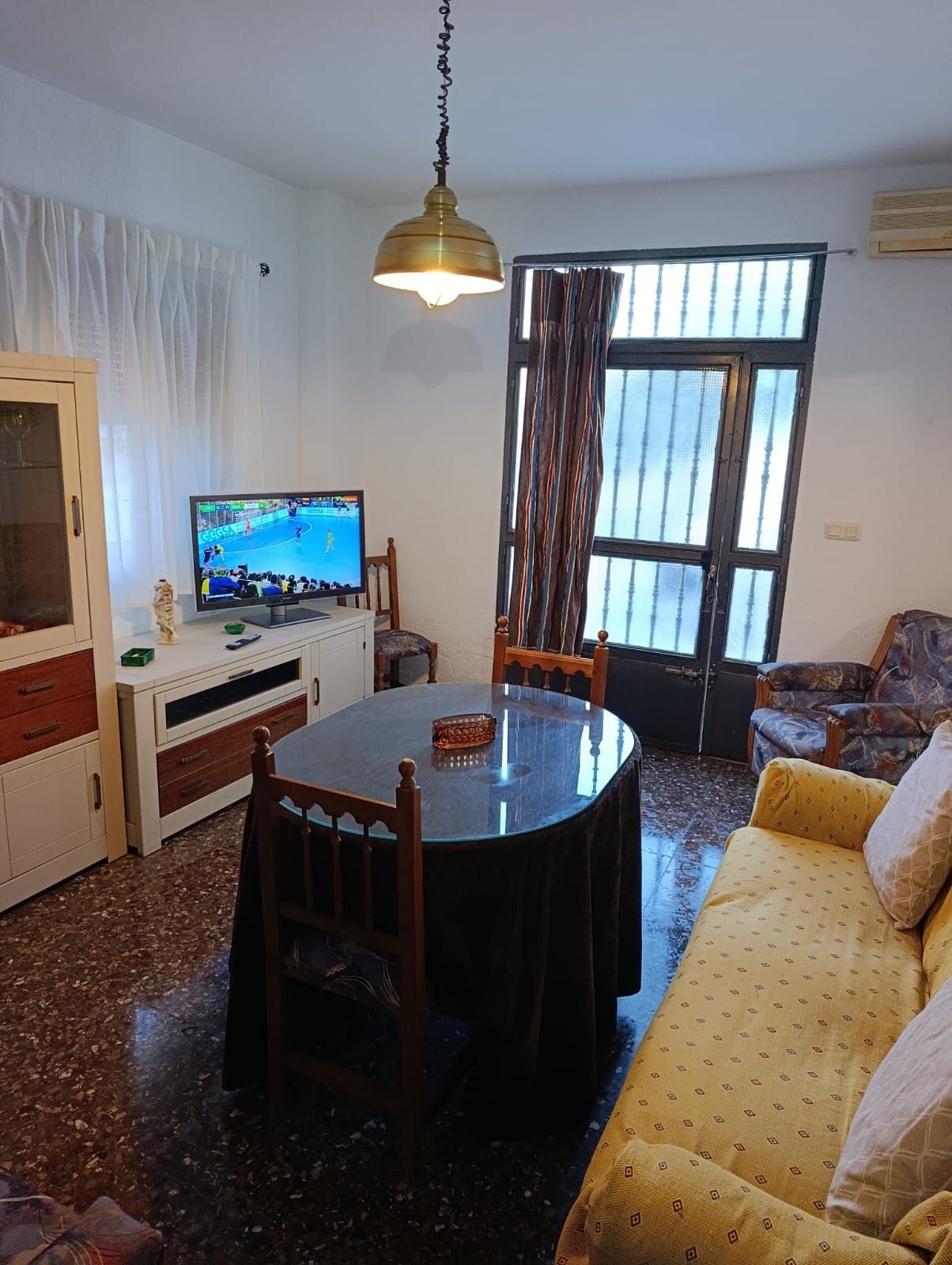 A living room with a round table, chairs, a television, a sofa, and a window with curtains.