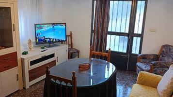 A living room with a round table, chairs, a television, a sofa, and a window with curtains.