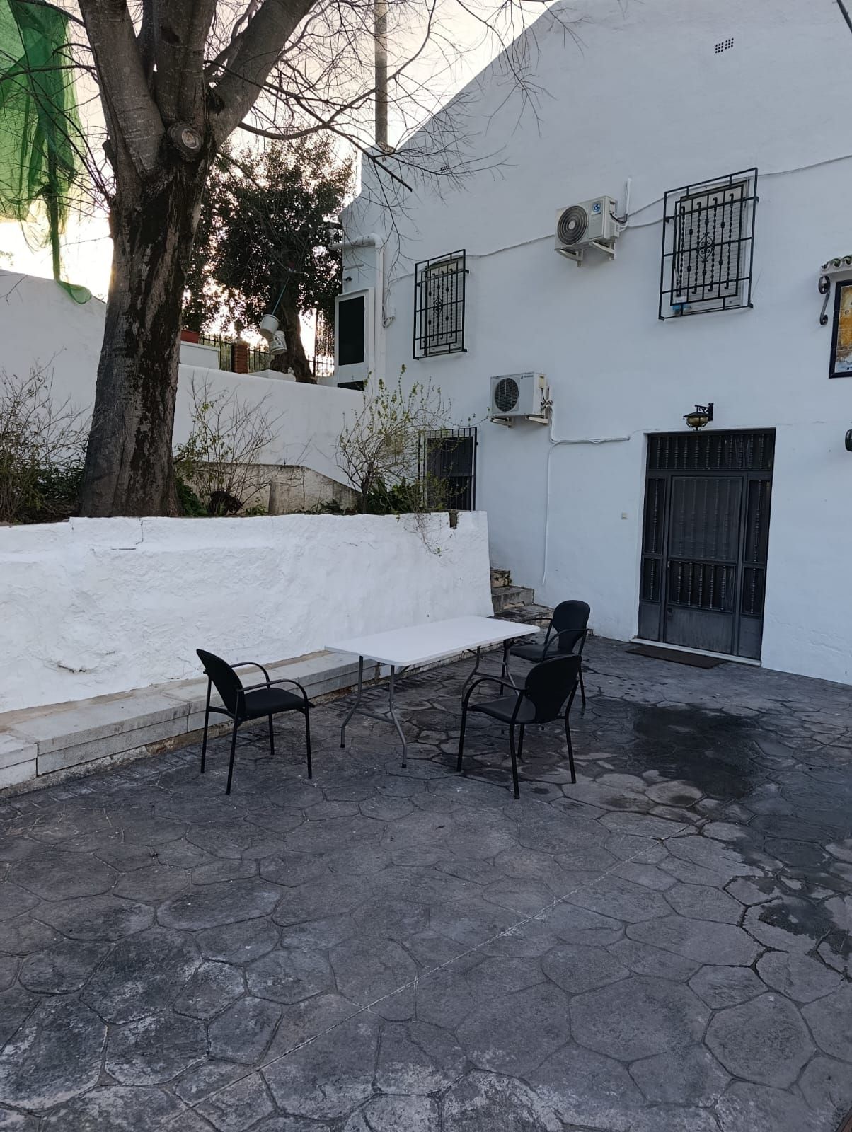 Un patio con un muro blanco, un árbol y una zona de estar con dos sillas y una mesa.