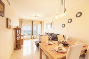 Un comedor con una mesa de madera dispuesta para el desayuno, un sofá y vistas al mar a través de amplias ventanas.