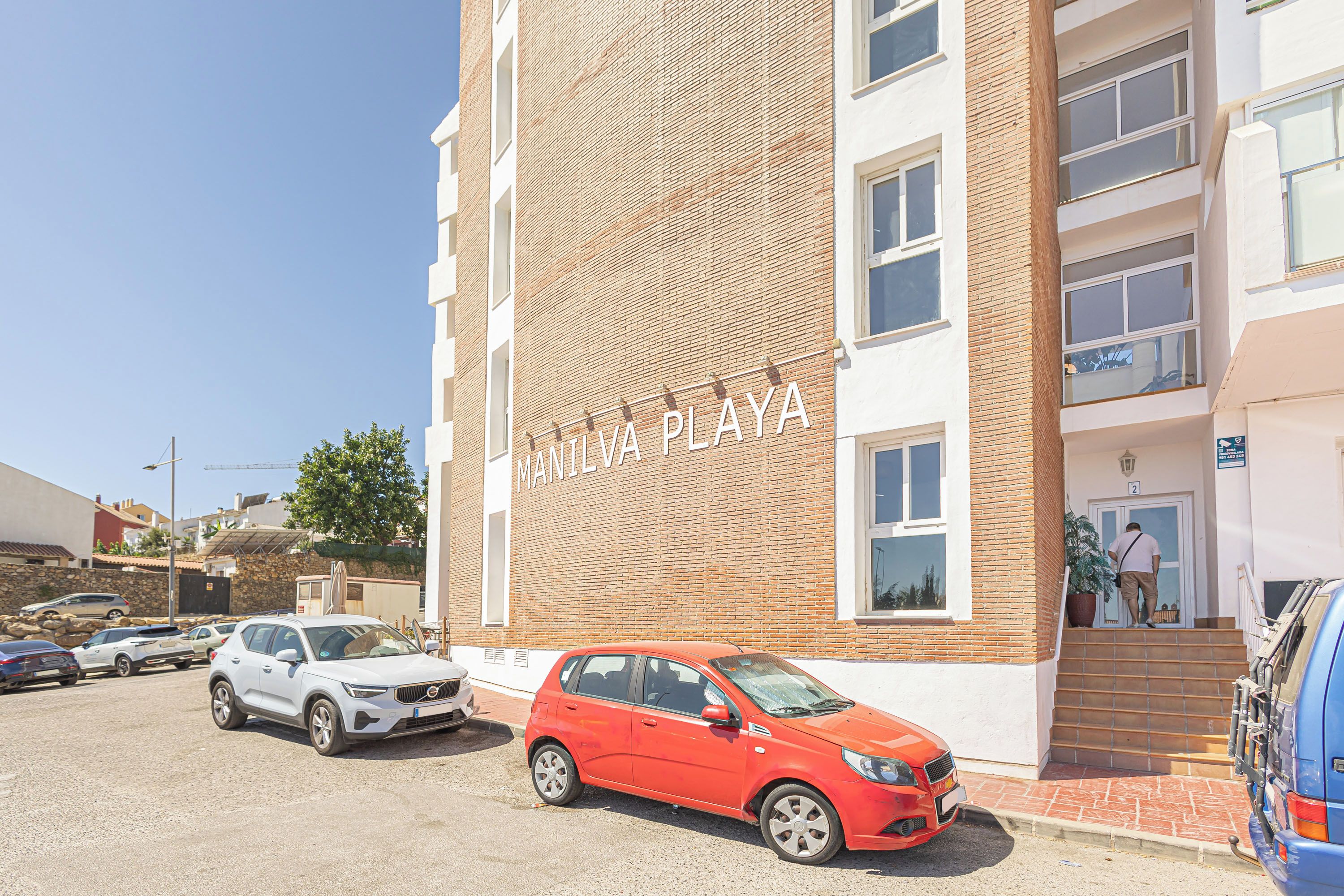 Un edificio con un letrero que dice 'MANILVA PLAZA' y un estacionamiento con varios autos.