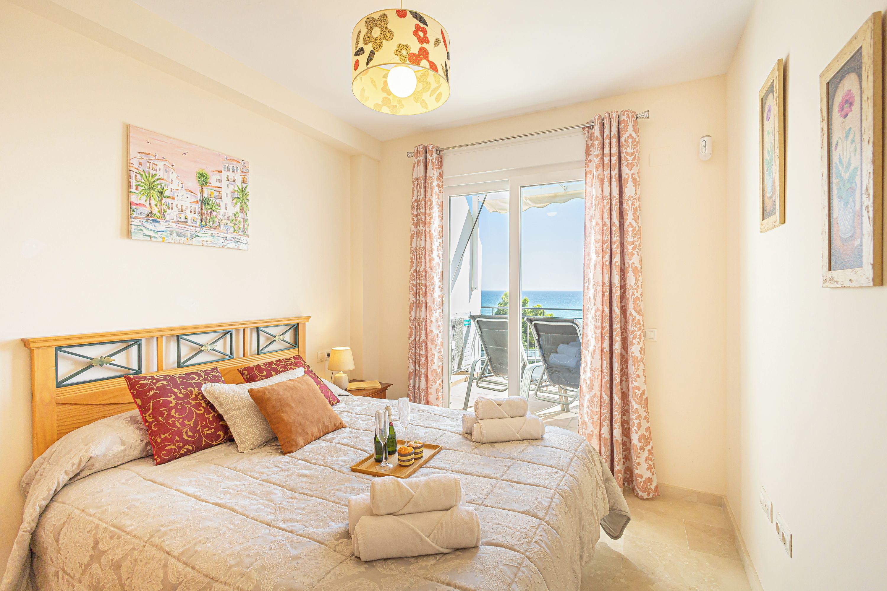 Habitación de hotel con cama, mesita de noche, ventana con cortinas, vista al mar y un cuadro en la pared.