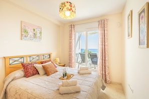 Habitación de hotel con cama, mesita de noche, ventana con cortinas, vista al mar y un cuadro en la pared.