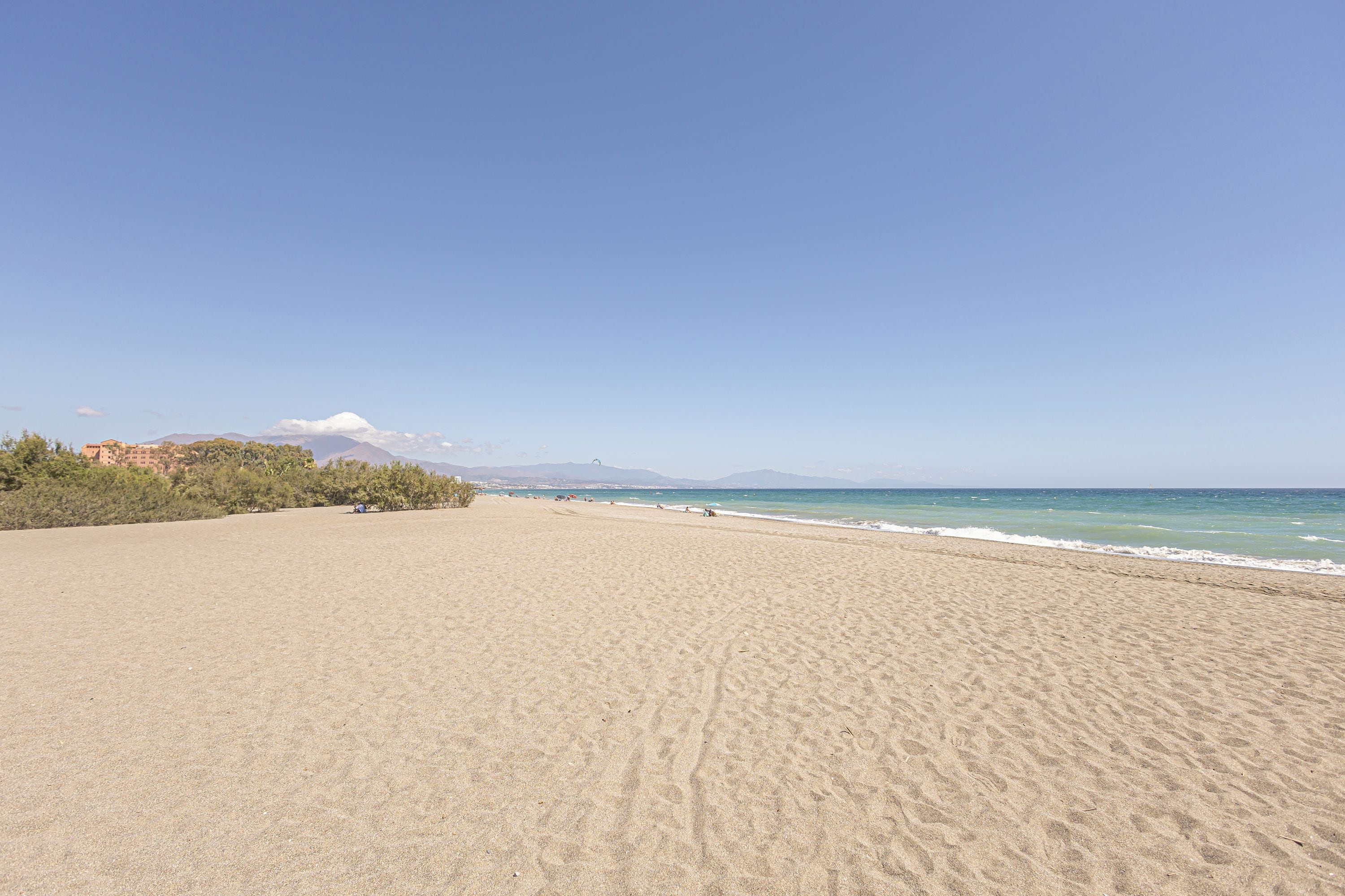 A wide sandy beach with a clear sky, a few people, and distant mountains.