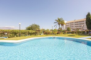 Une piscine aux eaux cristallines, bordée de chaises longues et d’ombrelles, entourée de verdure et d’un bâtiment d’hôtel.