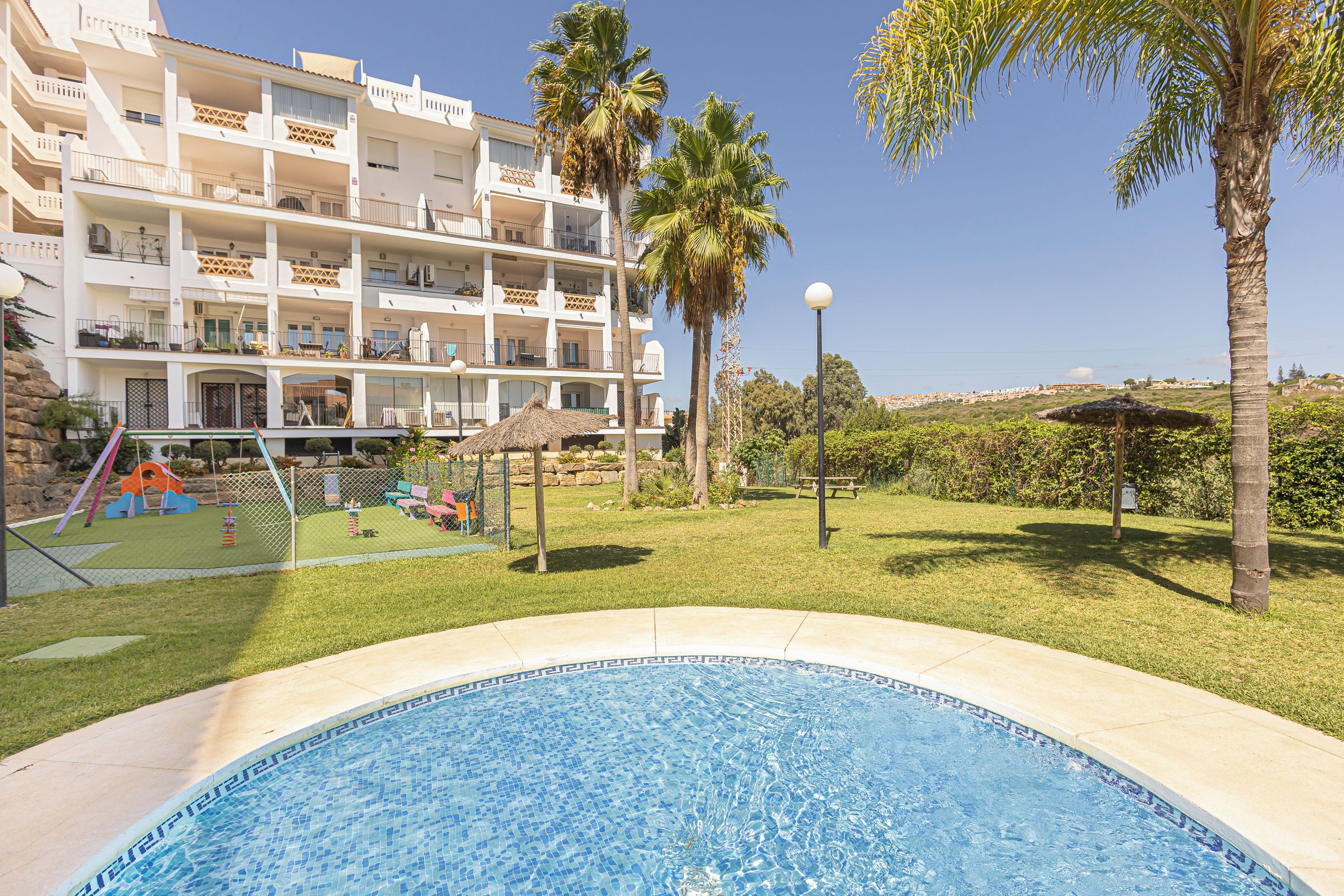 A swimming pool with a tiled edge, surrounded by a grassy area with palm trees and a playground.