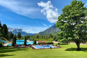 Ein Schwimmbeckenbereich mit Bergblick, umgeben von viel Grün und einem wolkenlosen Himmel.