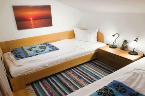 Ein Einzelbett mit Holz Kopfteil, weißer Bettwäsche, einem farbenfrohen Streifen Teppich, einem Nachttisch mit Lampe und einem Wandbild von einem Sonnenuntergang.