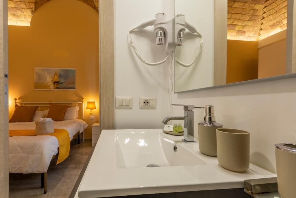 Una camera d'albergo con un grande specchio, un lavandino bianco con rubinetteria moderna e un letto con coperta gialla.