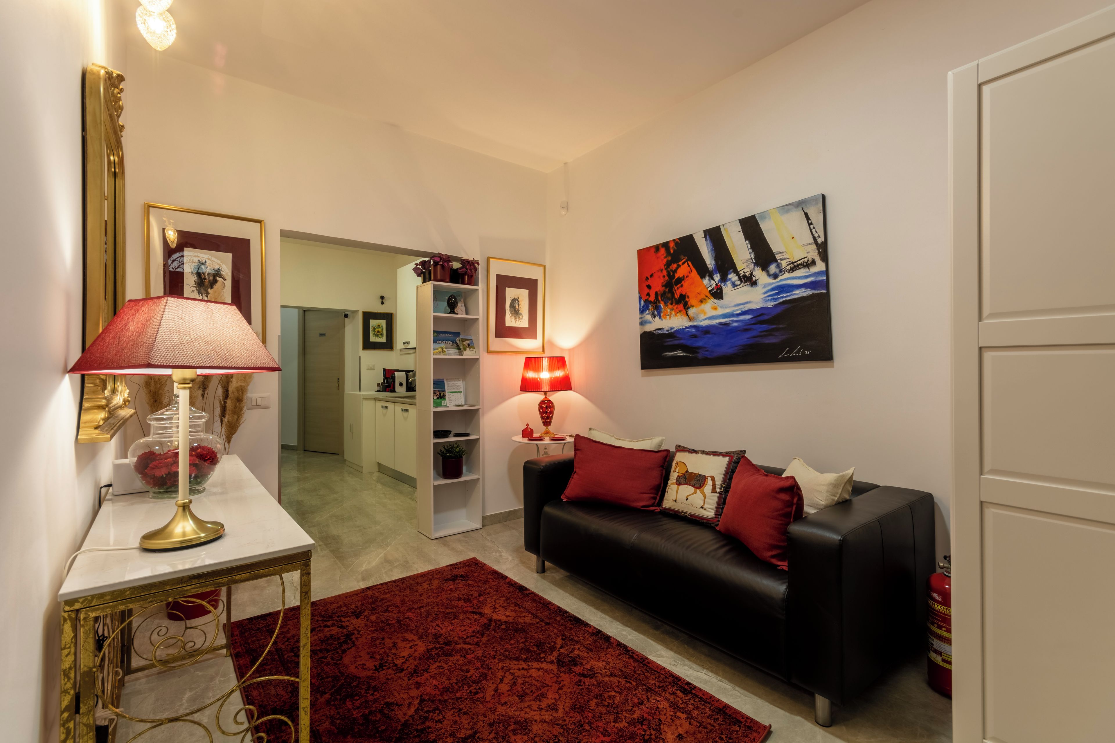 A living room with a black leather sofa, red cushions, a red rug, a gold-framed side table, a red lamp, and a painting on the wall.