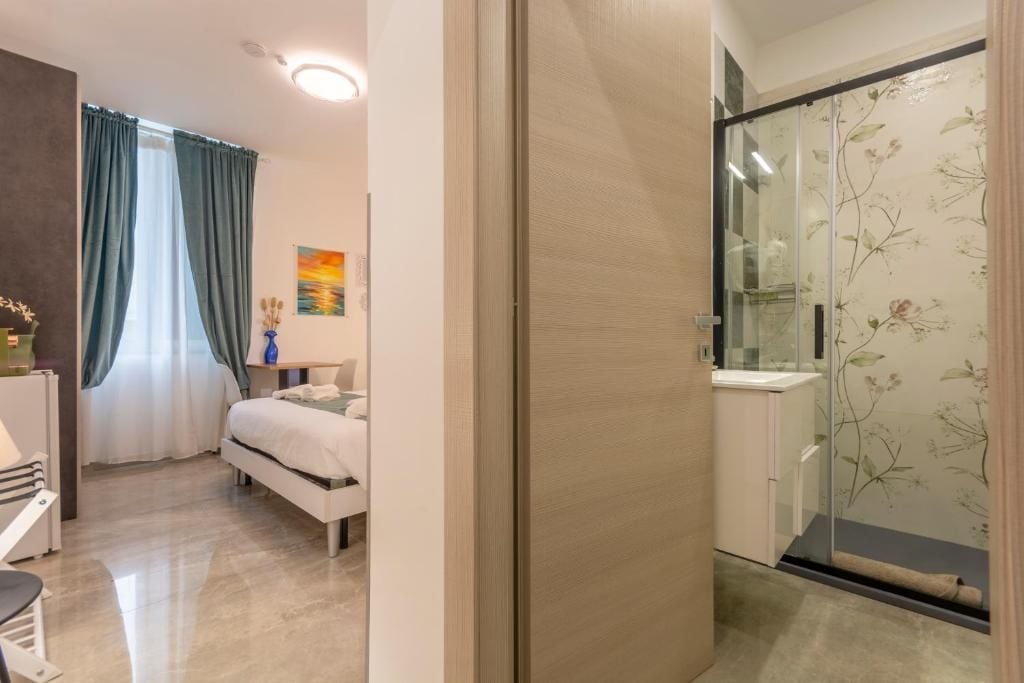 Un dormitorio con cama, escritorio y silla, un baño con cabina de ducha de vidrio, y vistas al exterior.