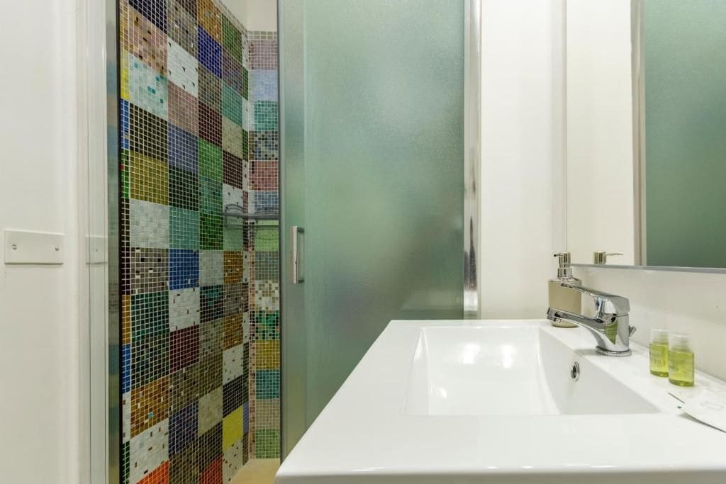 Un baño con lavamanos blanco, espejo y ducha con pared de mosaicos de colores.