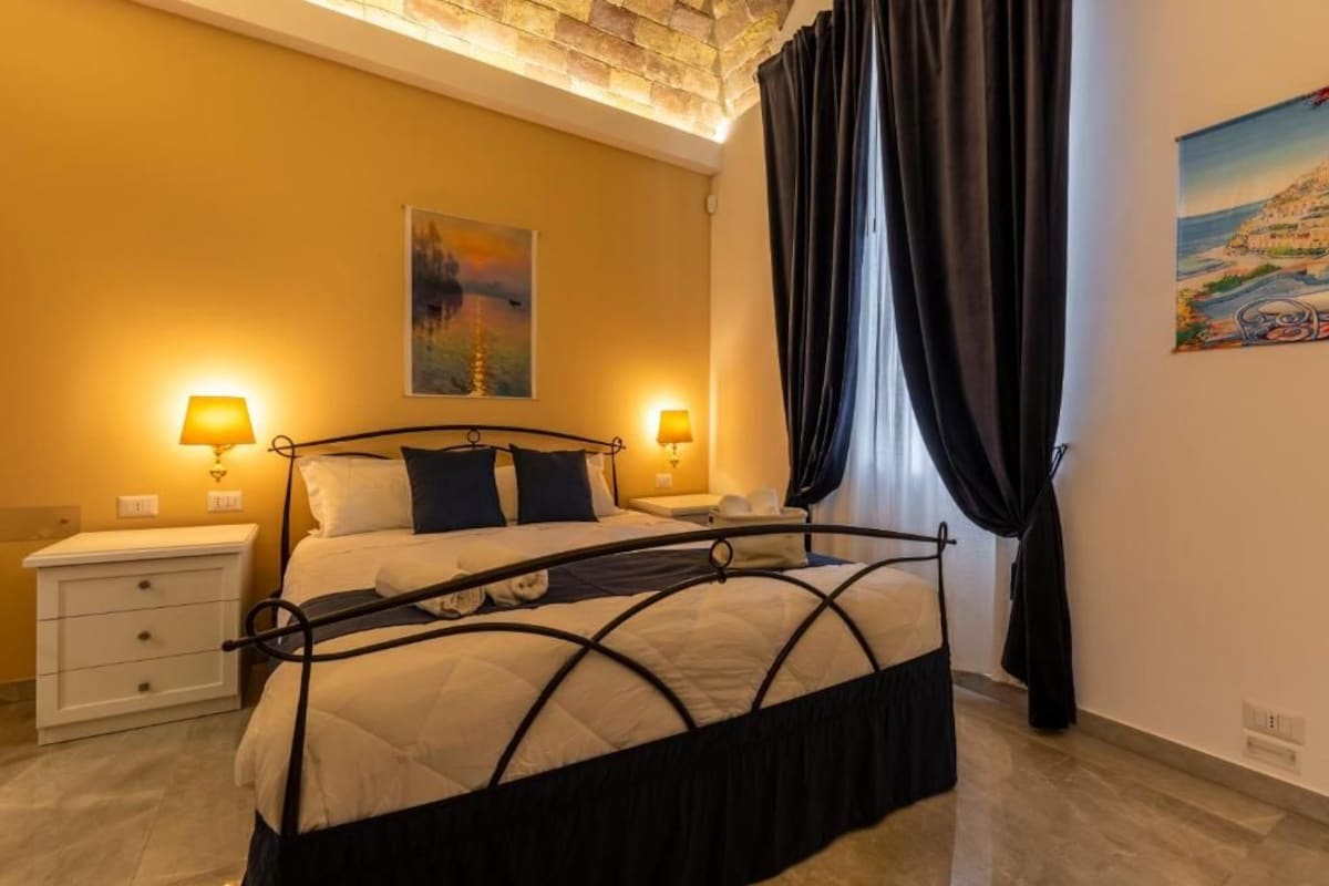 A bedroom with a bed, two bedside tables, a painting on the wall, and a window with curtains.