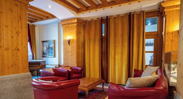 Ein gemütliches Zimmer mit Holzbalken, einem roten Ledersofa, einem Holztisch und einem großen Fenster mit Vorhängen.