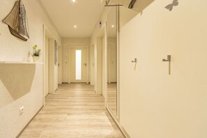 A hallway with a wooden floor, a white wall with a shelf holding a plant, and a door at the end. - Holiday apartment with 2 bedrooms (Norddeich)