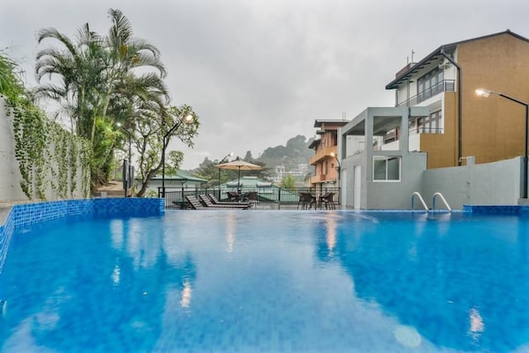 A swimming pool with a clear blue water surface, surrounded by a fence, and adjacent to a building with a balcony. - Hotel TravllersNest Kandy (Kandy)