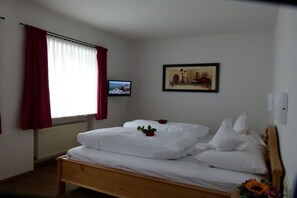 Un dormitori amb un llit de fusta, roba de llit blanca, una cortina vermella, un quadre amb marc a la paret i un televisor a l'esquerra.