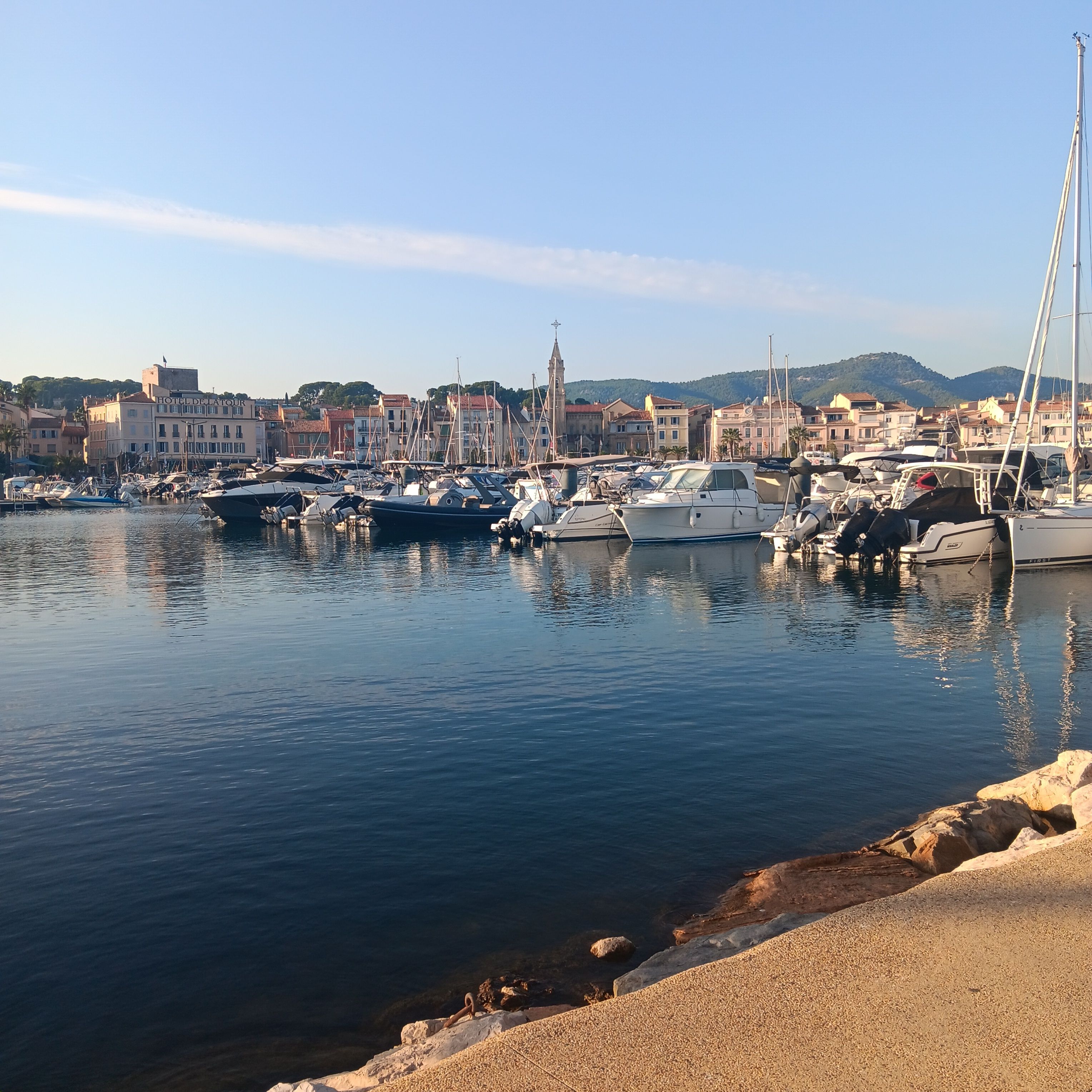 A marina with boats docked, a coastal town in the background, and a prominent building labeled 'HOTEL DELLA TOUR'.