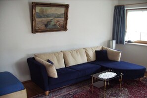Een woonkamer met een donkerblauwe bank, een klein rond salontafeltje, een kamerkleed met een patroon, een lijstwerk met een schilderij aan de muur, en een raam met gordijnen.