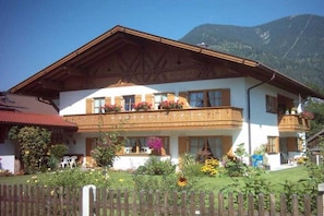 A two-story building with a balcony, surrounded by a wooden fence, and a mountain in the background. - Holiday apartment with 1 bedroom (Garmisch-Partenkirchen)
