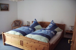 A wooden bed with a blue and white patterned bedspread and pillows, a wooden nightstand with a lamp, and a chair in a room with a plain wall and a door. - Holiday apartment with 1 bedroom (Garmisch-Partenkirchen)