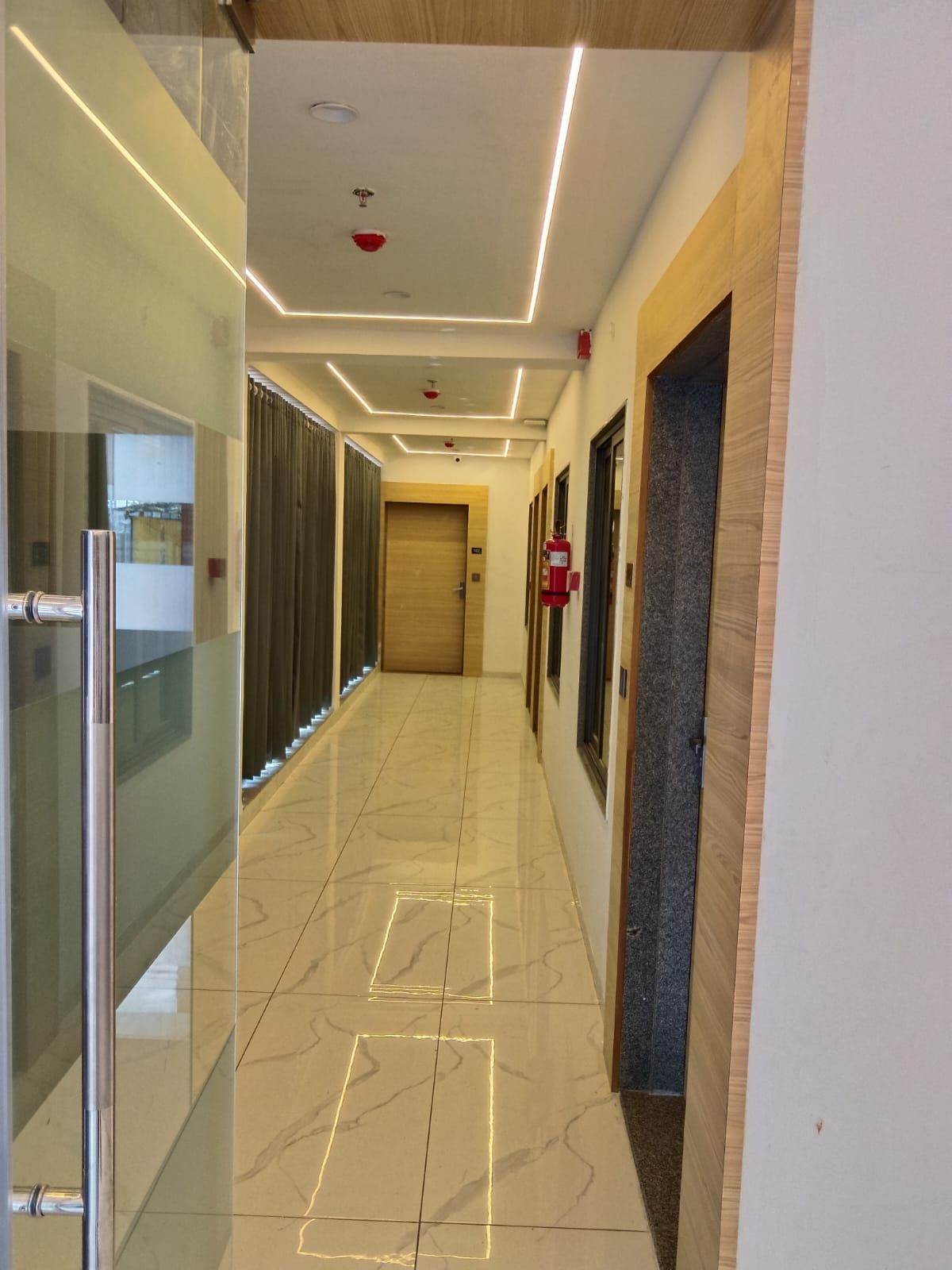 A corridor with a glass door on the left, a fire extinguisher on the right, and a wooden door at the end.