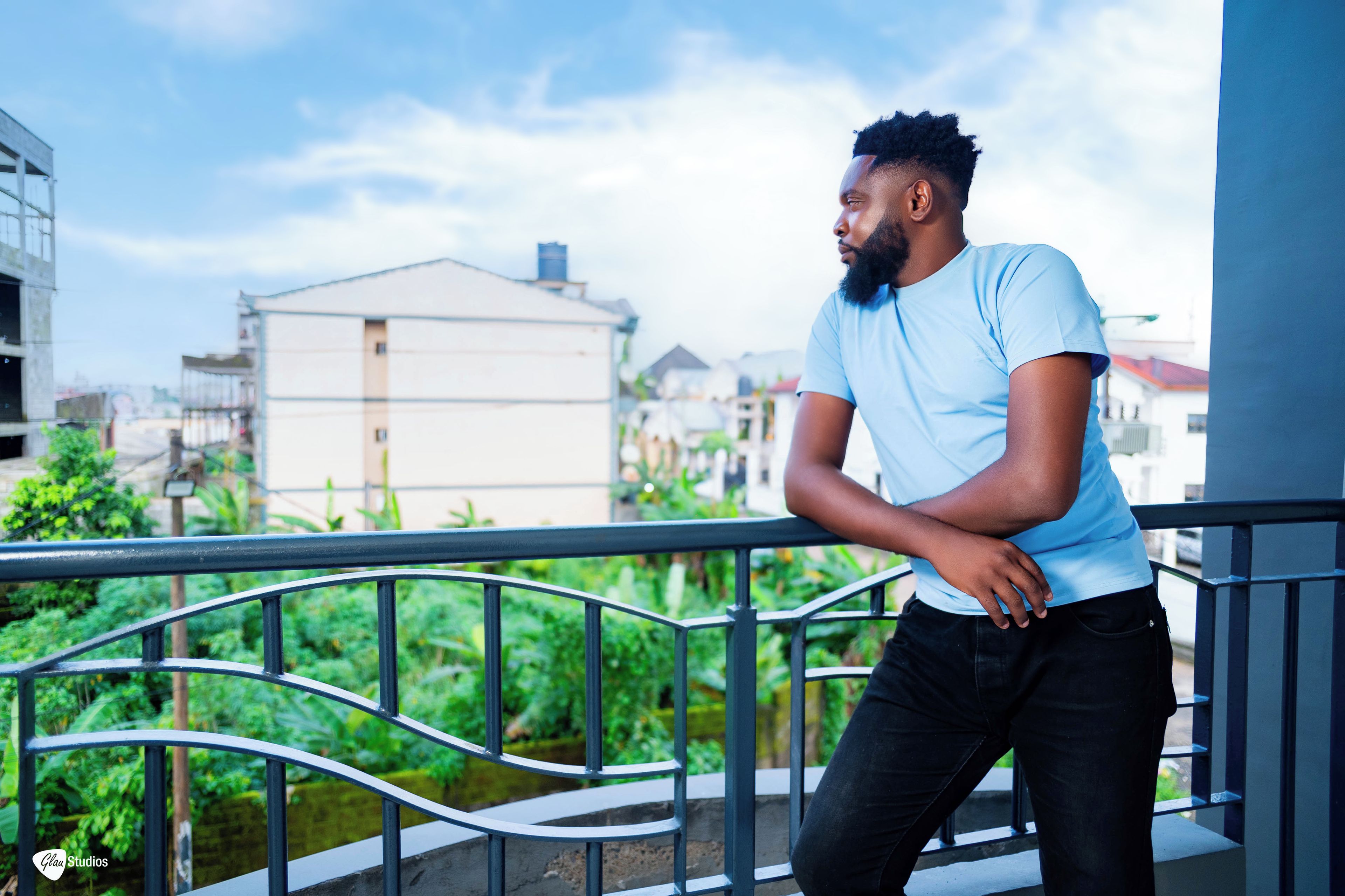 A person leaning on a balcony railing with a view of buildings and greenery in the background.