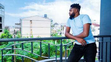 A person leaning on a balcony railing with a view of buildings and greenery in the background.