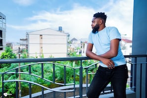 A person leaning on a balcony railing with a view of buildings and greenery in the background. - Residence SOLEN (Douala)