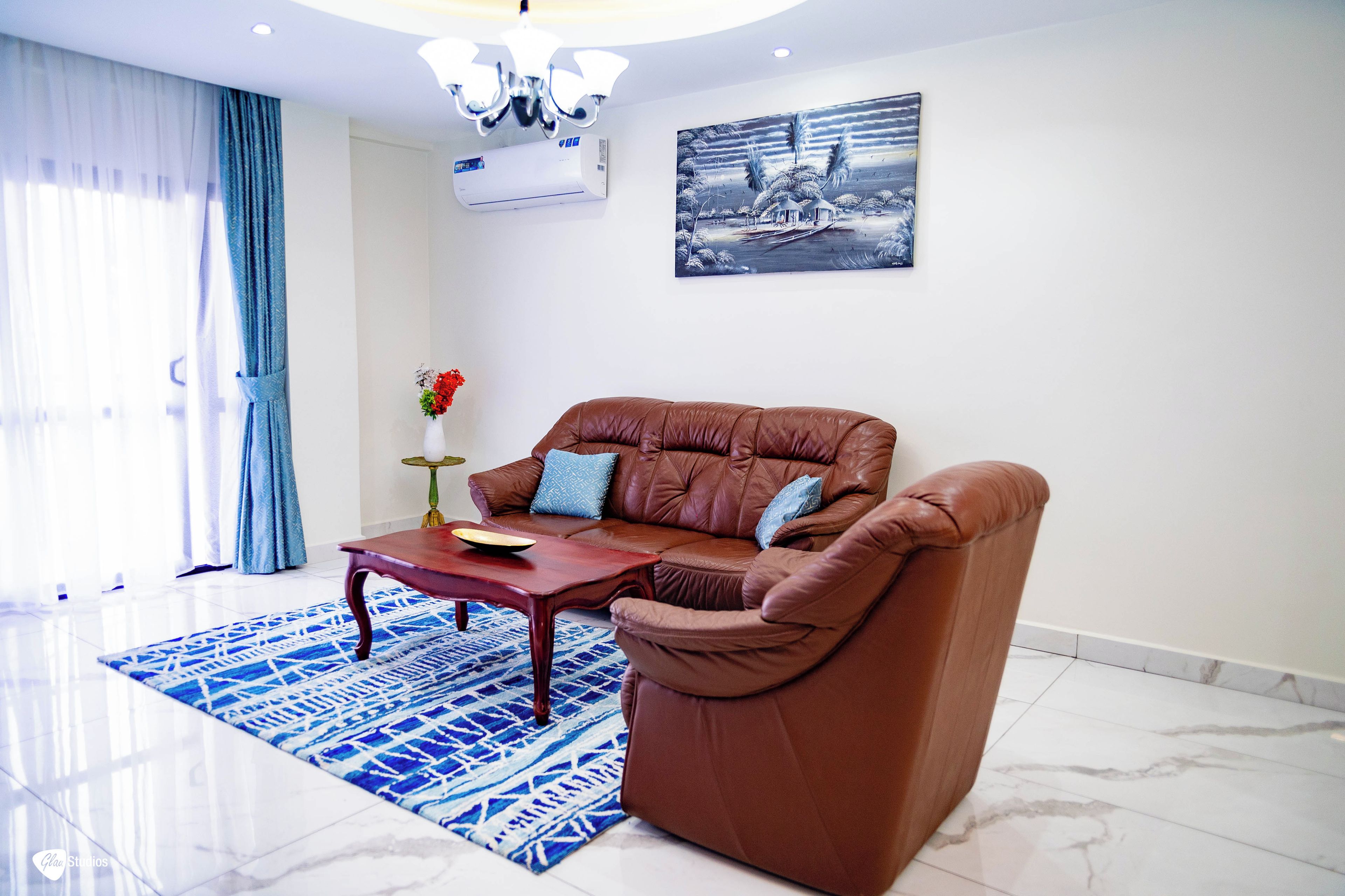 A living room with a brown leather sofa, a wooden coffee table, a blue patterned rug, a vase with flowers, and a painting on the wall.