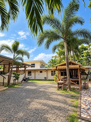 A tropical resort with a thatched-roof hut, palm trees, and a gravel courtyard.
