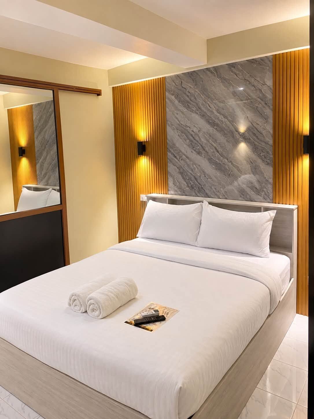 Une chambre d’hôtel moderne avec un grand lit, une tête de lit en bois, un mur décoratif en marbre et un miroir.