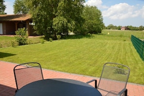 Eine Terrasse mit Tisch und Stühlen, ein gepflegter Rasen und im Hintergrund ein Gebäude mit rotem Dach.