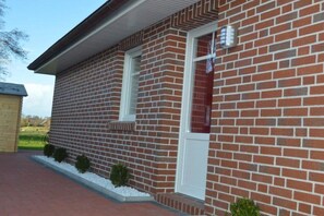 Un bâtiment en briques avec une porte et une fenêtre blanches, entouré d’un petit jardin agrémenté de gravier blanc et d’arbustes verts.
