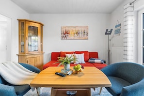 Ein modernes Wohnzimmer mit einem roten Sofa, einem Holzschrank, einem Gemälde und einem Tisch mit Pflanze und Obstschale.