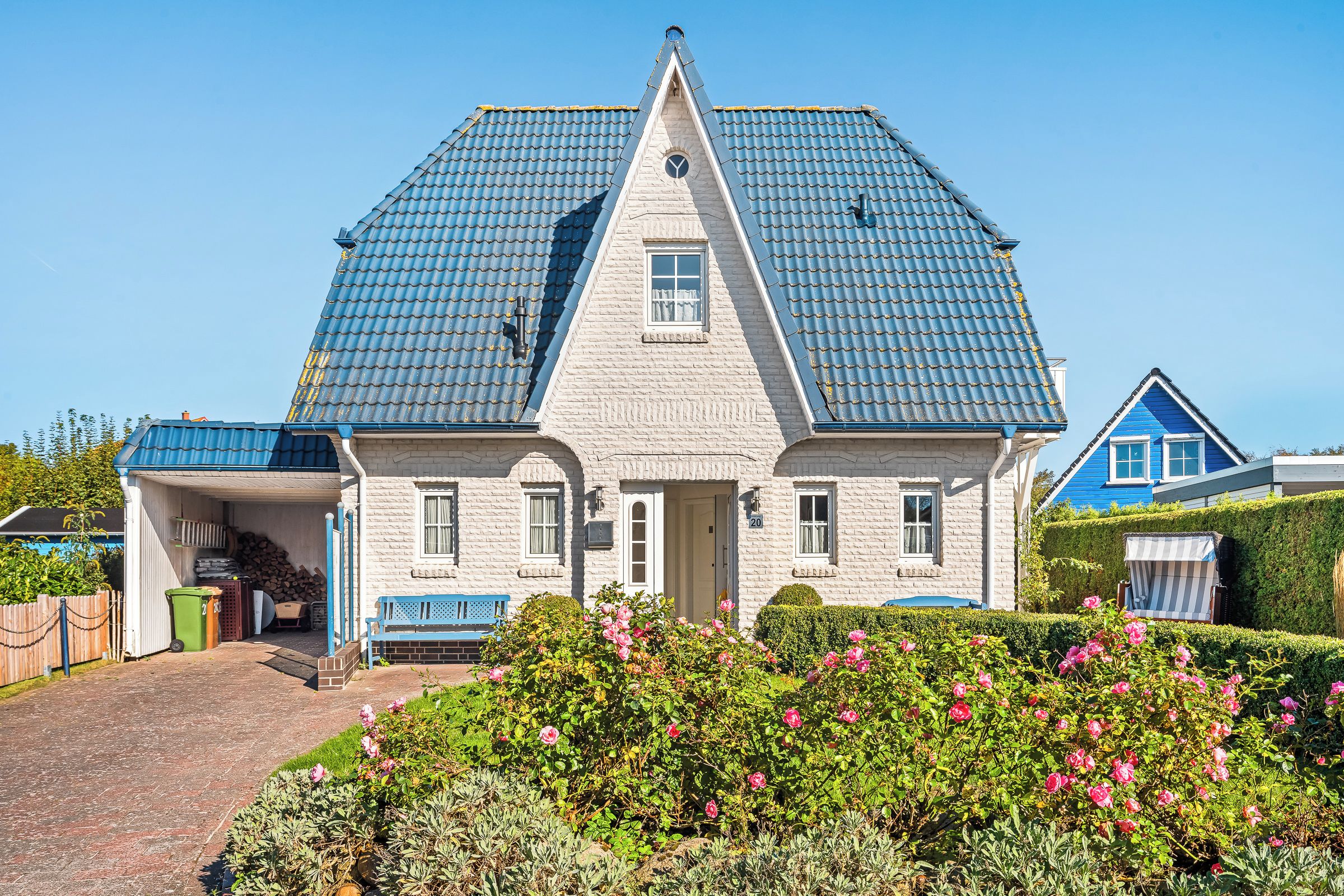 A two-story house with a steeply pitched roof, a front porch, and a well-maintained garden with pink flowers.