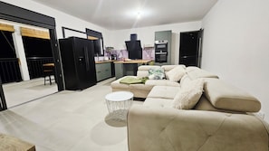 Una sala de estar moderna con un sofá color beige, una mesita redonda y una cocina al fondo.