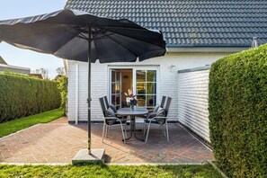 Eine Terrasse mit Tisch und Stühlen, einem großen Sonnenschirm und einem weißen Gebäude mit einem Ziegeldach.