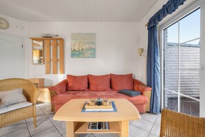 Una sala de estar con un sofá rojo, una mesa de madera con una bandeja, sillas de mimbre, un cuadro y un ventanal con cortinas.