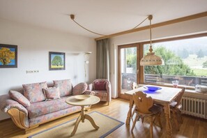 A cozy living room with a sofa, a dining table with chairs, a hanging lamp, and a view through a large window. - Holiday apartment with 1 bedroom (Farchant)