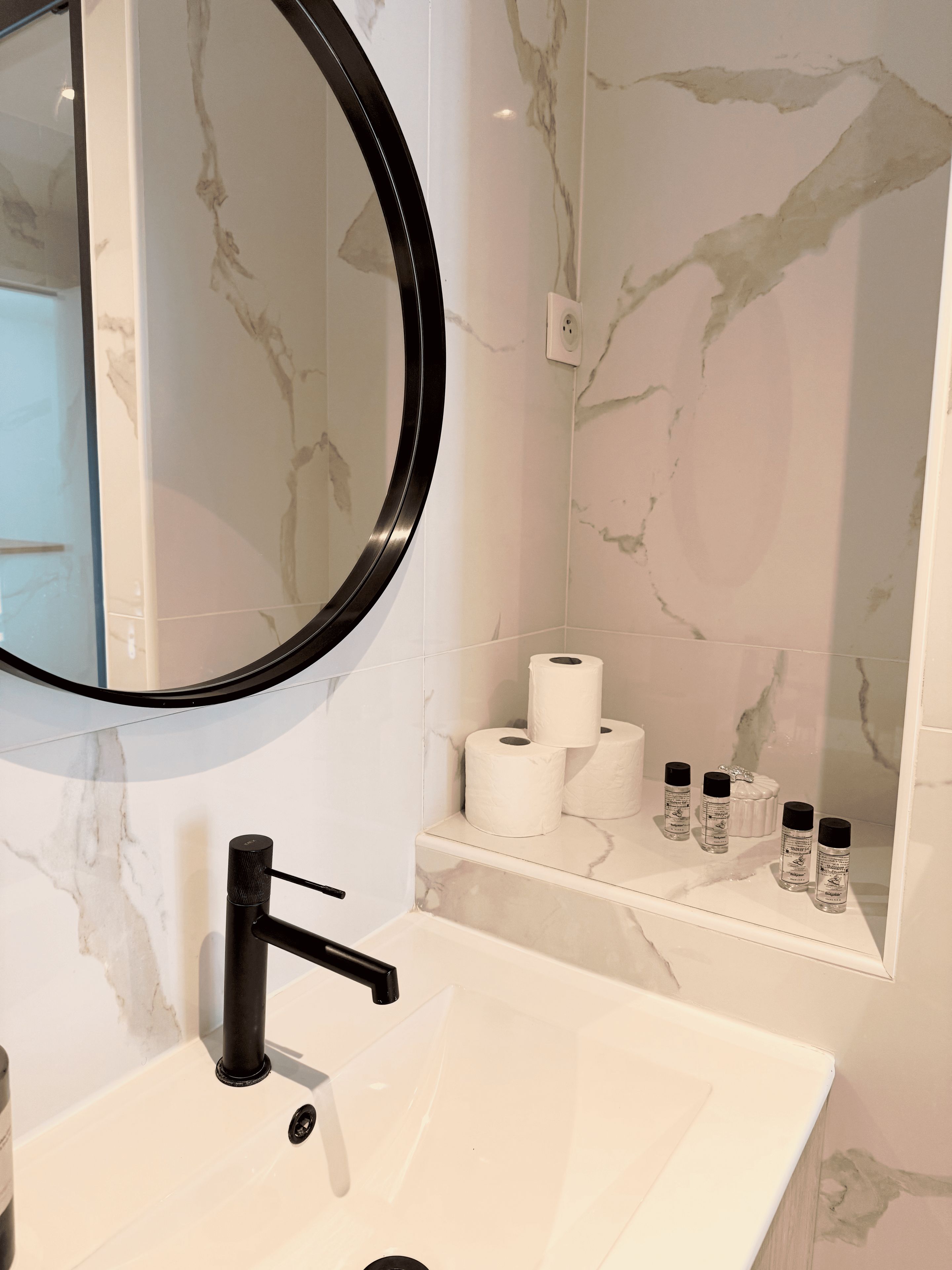 A bathroom with a white sink, black faucet, and a mirror with a black frame. There are two rolls of toilet paper and four bottles of hand soap on a shelf.