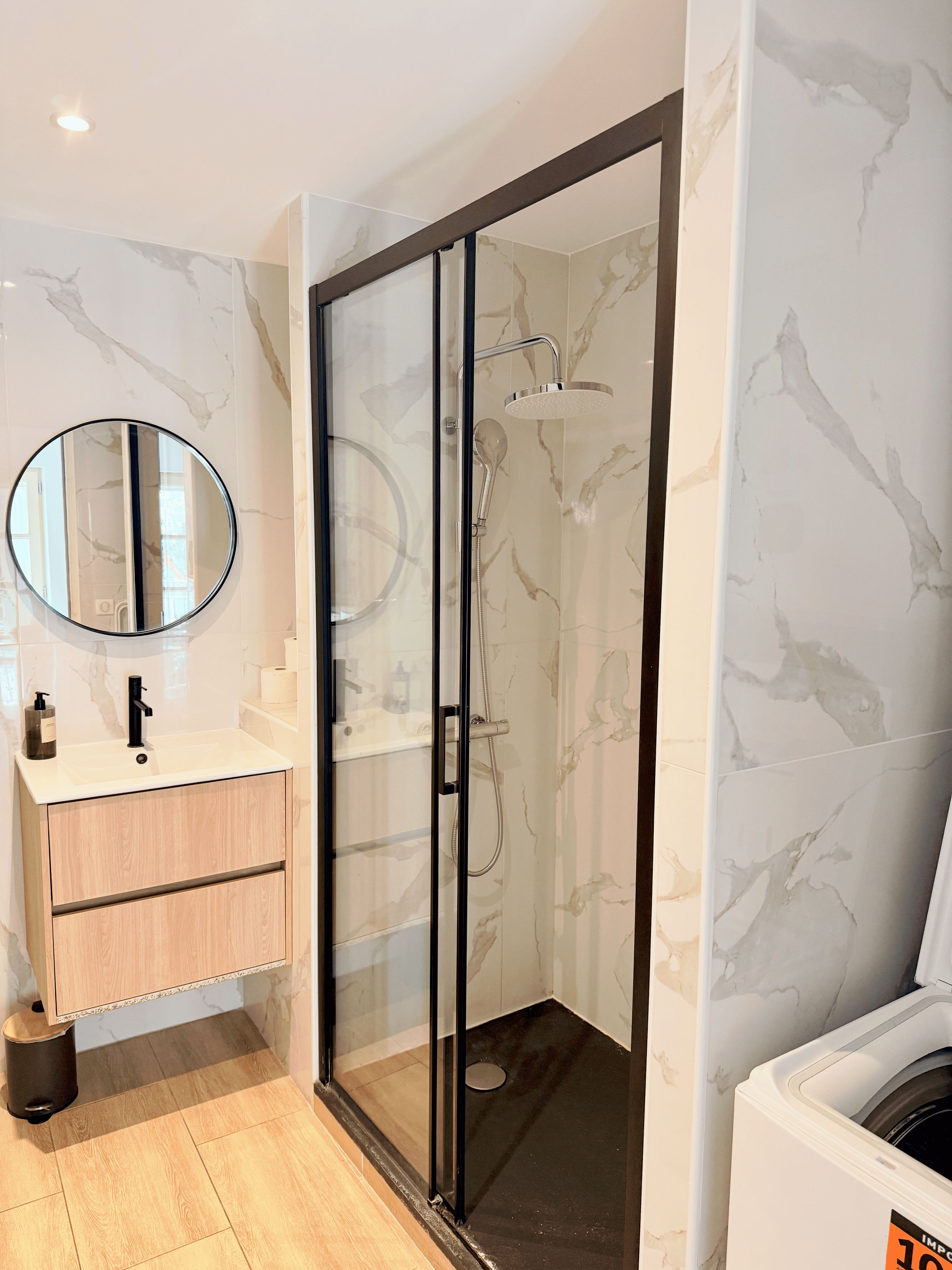 Une salle de bain moderne avec une douche à parois de verre, un miroir rond, un lavabo avec un distributeur de savon et des toilettes.