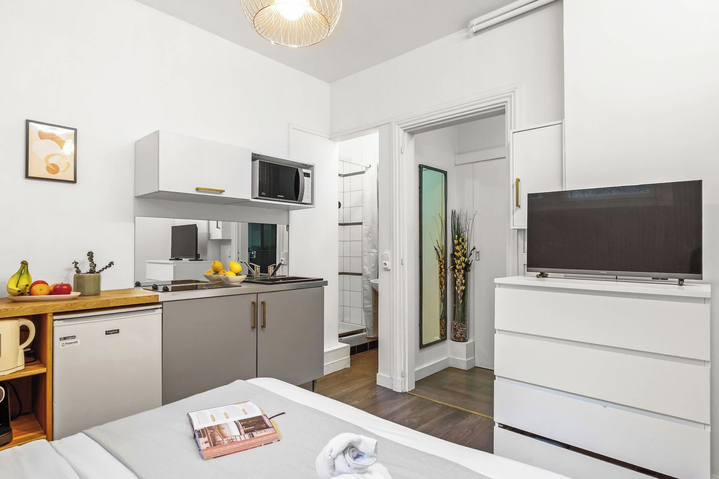 Een kleine hotelkamer met kitchenette, een bed, een televisie en een deur naar een badkamer.